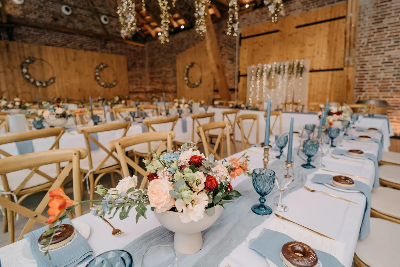 Festlich gedeckte Hochzeitstafel in einer Scheune mit blauen Gläsern, Blumen in Pfirsichtönen und Holzstühlen