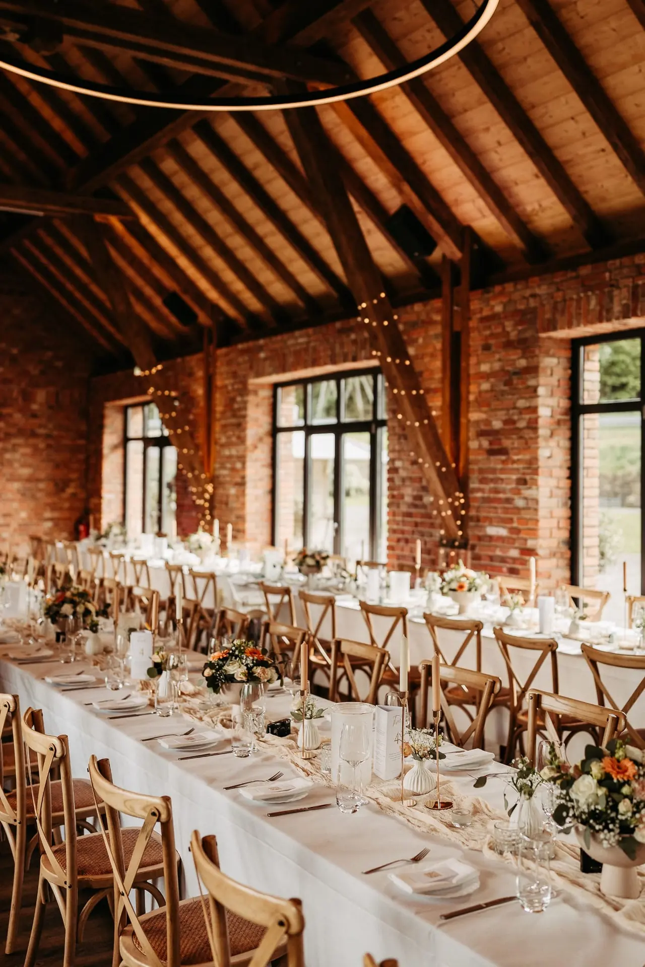 Hochzeit in einer Scheune mit langer weißer Tafel, Lichterketten und Holzstühlen